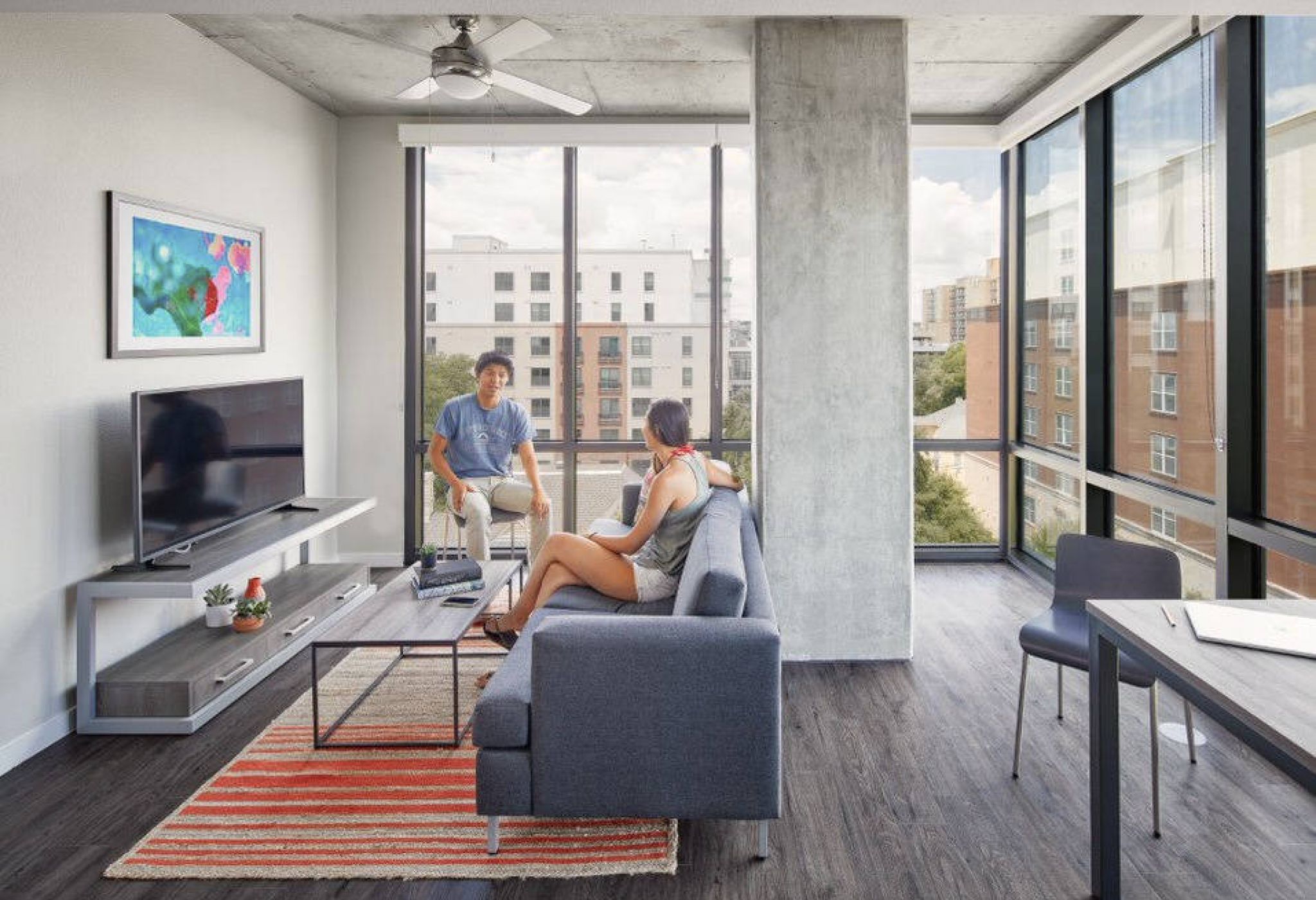 Two people talk in a modern, bright apartment with large windows, a sofa, TV, and city views.
