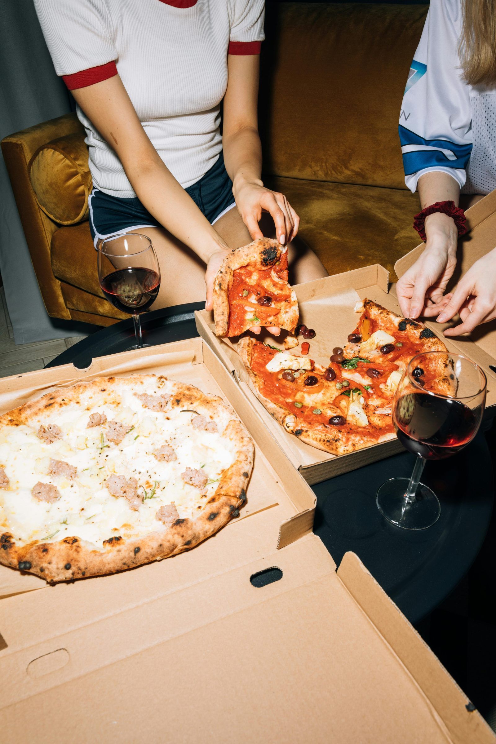 Two people share pizza from boxes on a table with two glasses of red wine, sitting on a couch.