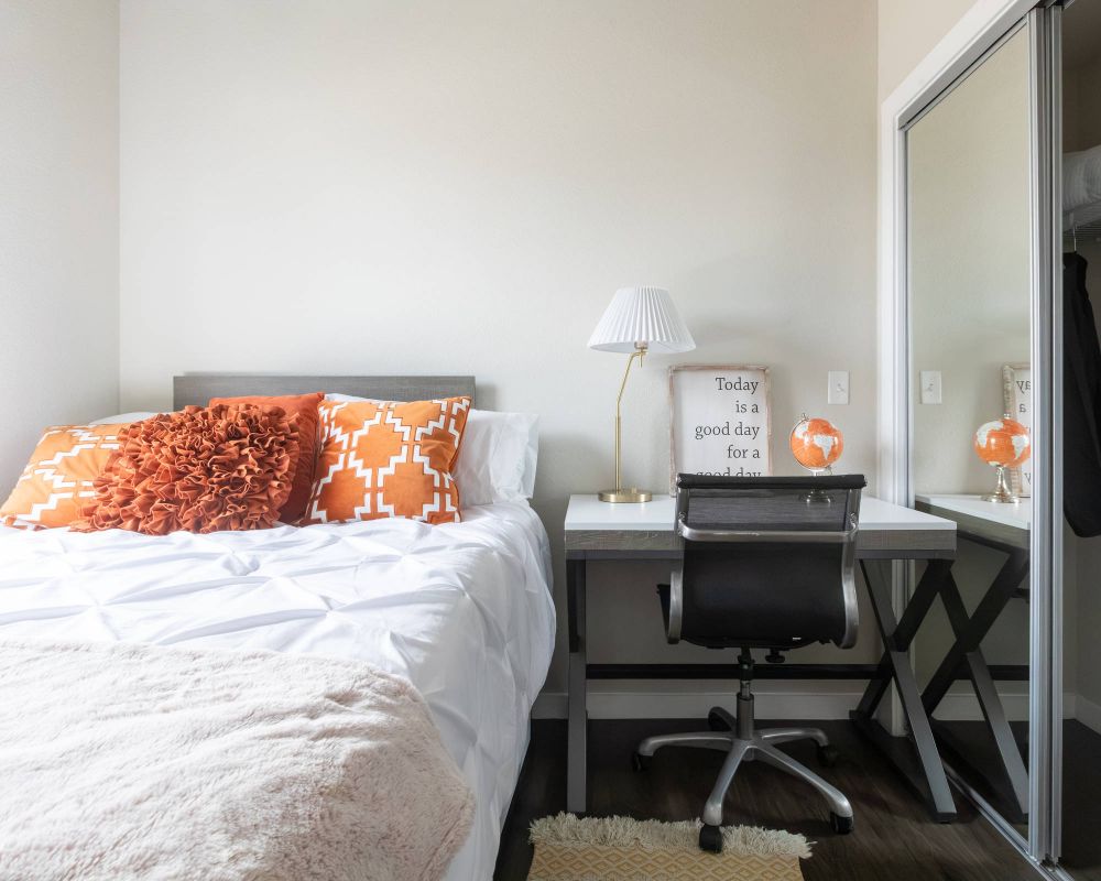 A tidy bedroom with a bed, orange pillows, a desk, chair, lamp, globe, and a mirrored closet.