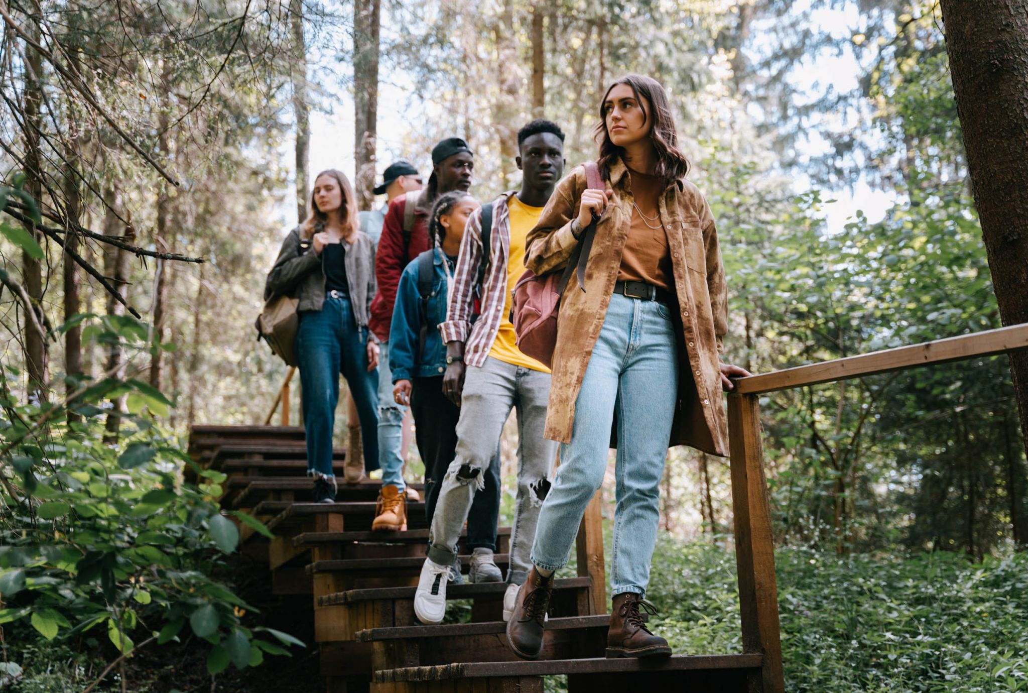 A group of young adults hike down wooden stairs in a forest, surrounded by lush green trees and sunlight.