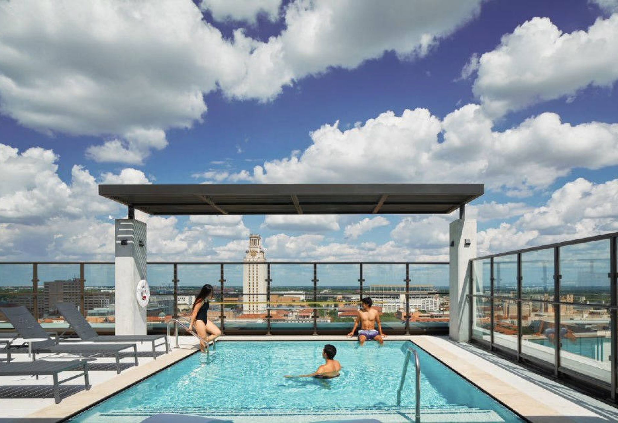 Three people relax in a rooftop pool under a pergola, with city skyline and blue sky with clouds in the background.