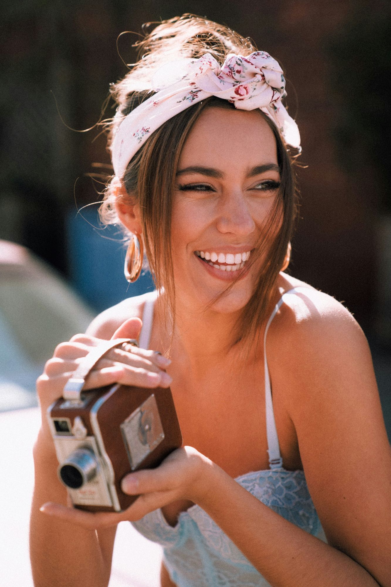 Smiling woman with a floral headscarf holds a vintage camera outdoors, sunlight illuminating her face.
