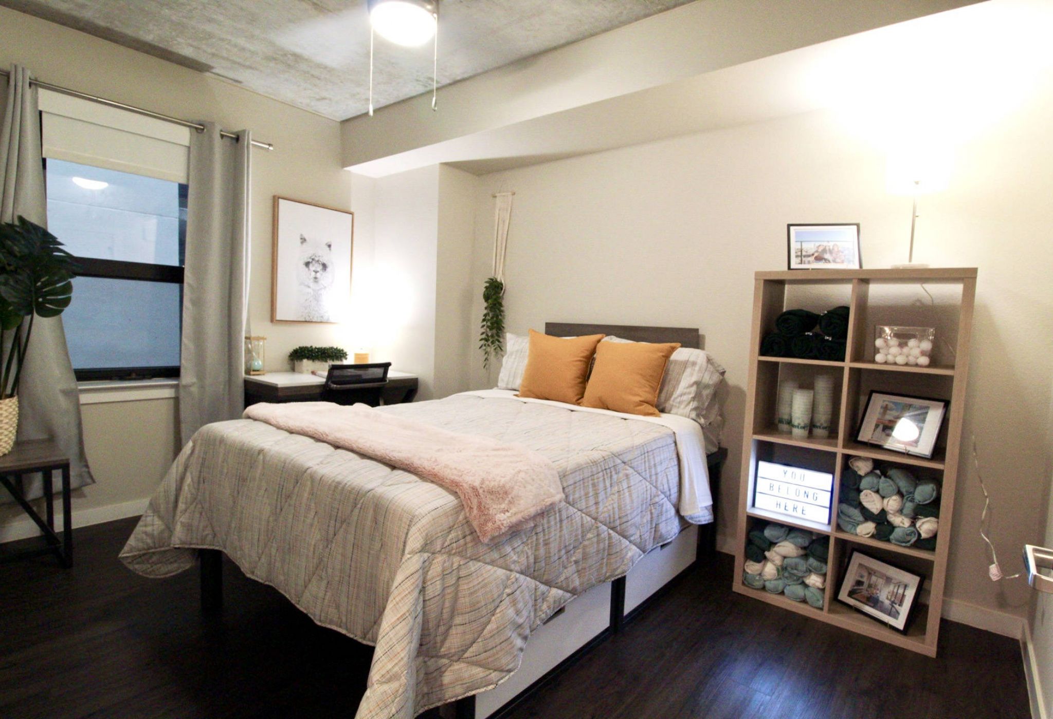 Cozy bedroom with a bed, desk by the window, and shelves with towels, decor, and a lightbox sign.