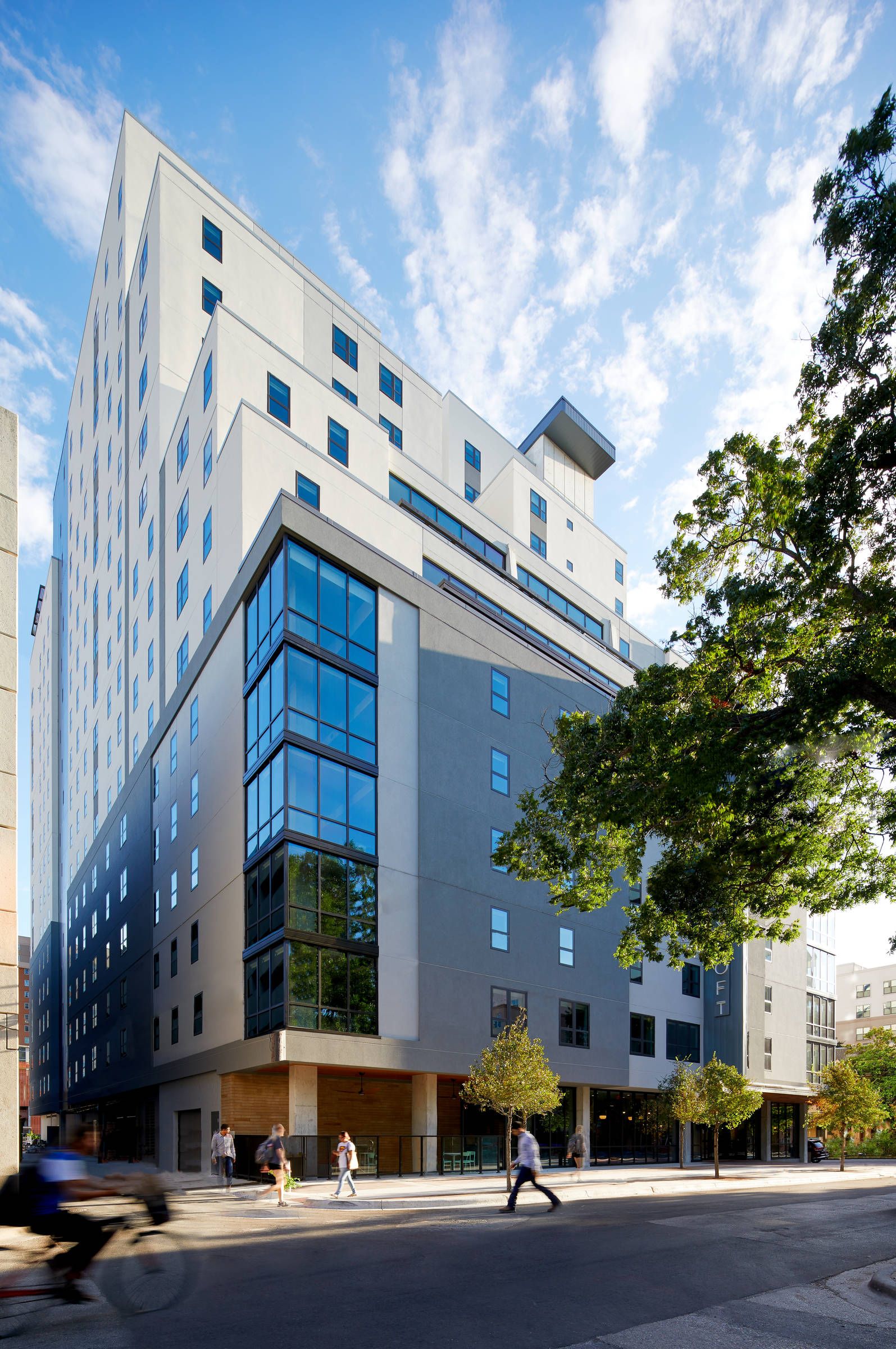 Modern multi-story building with large windows, people walking on the sidewalk, and a tree in the foreground.