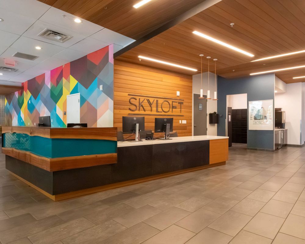Modern lobby with a colorful geometric accent wall, front desk, computers, and "SKYLOFT" sign on wood-paneled wall.