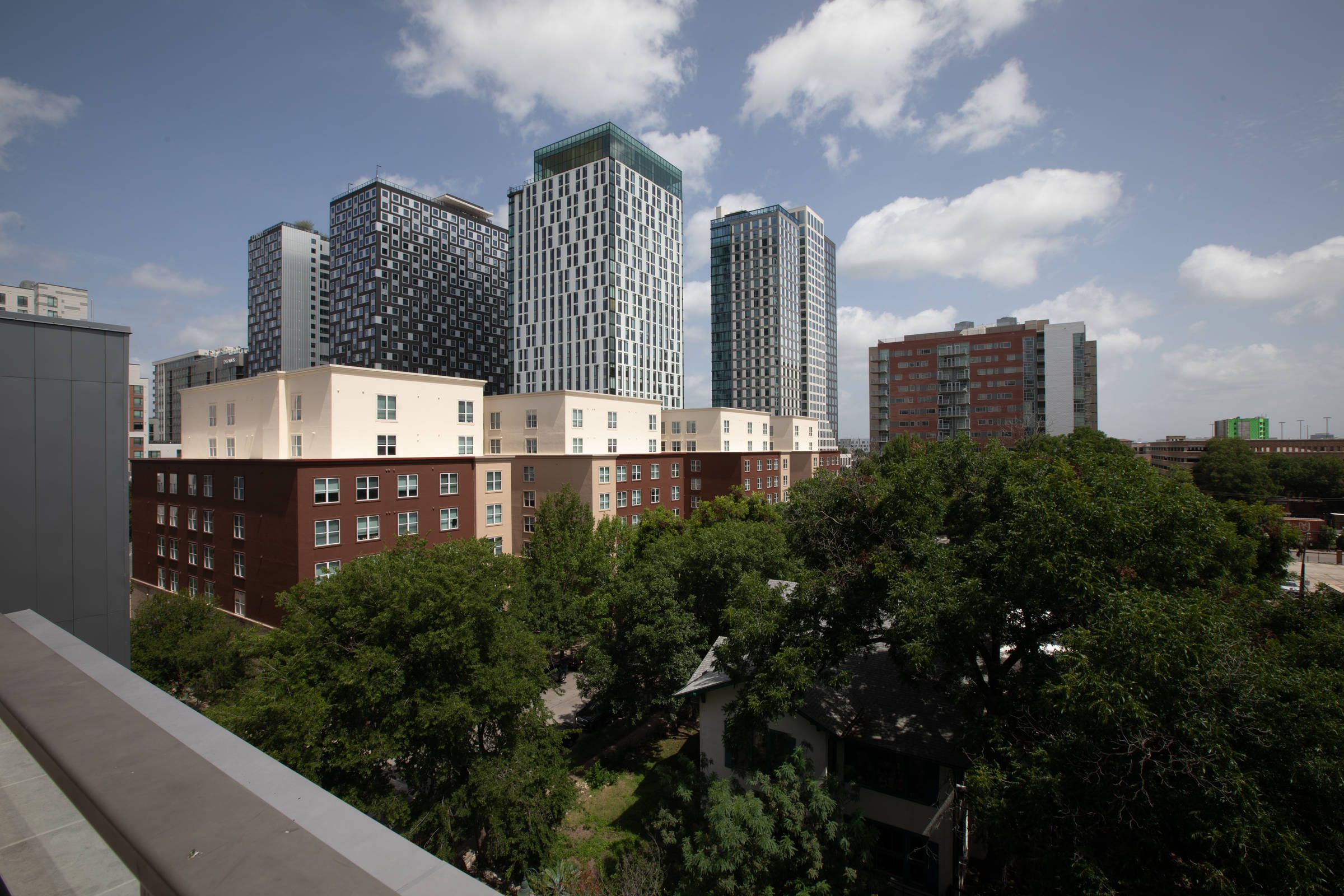 Tall modern buildings tower over shorter brick apartments and green trees under a partly cloudy sky.