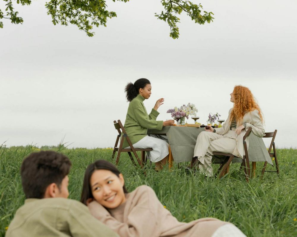 Two women sit at a picnic table while a man and woman relax on the grass, all enjoying a peaceful outdoor setting under a tree.