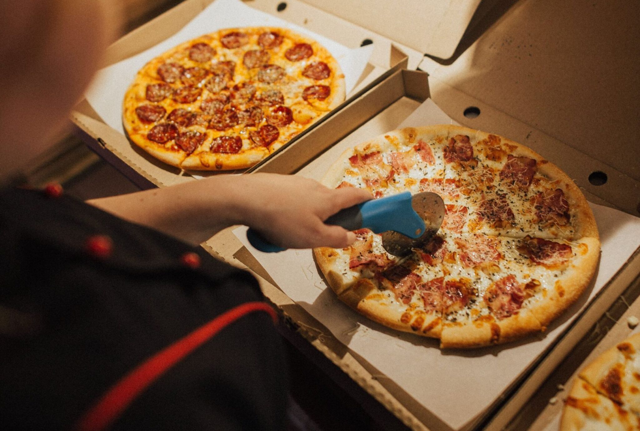 Person using a pizza cutter to slice a pizza in a box, with two more pizzas in boxes nearby.