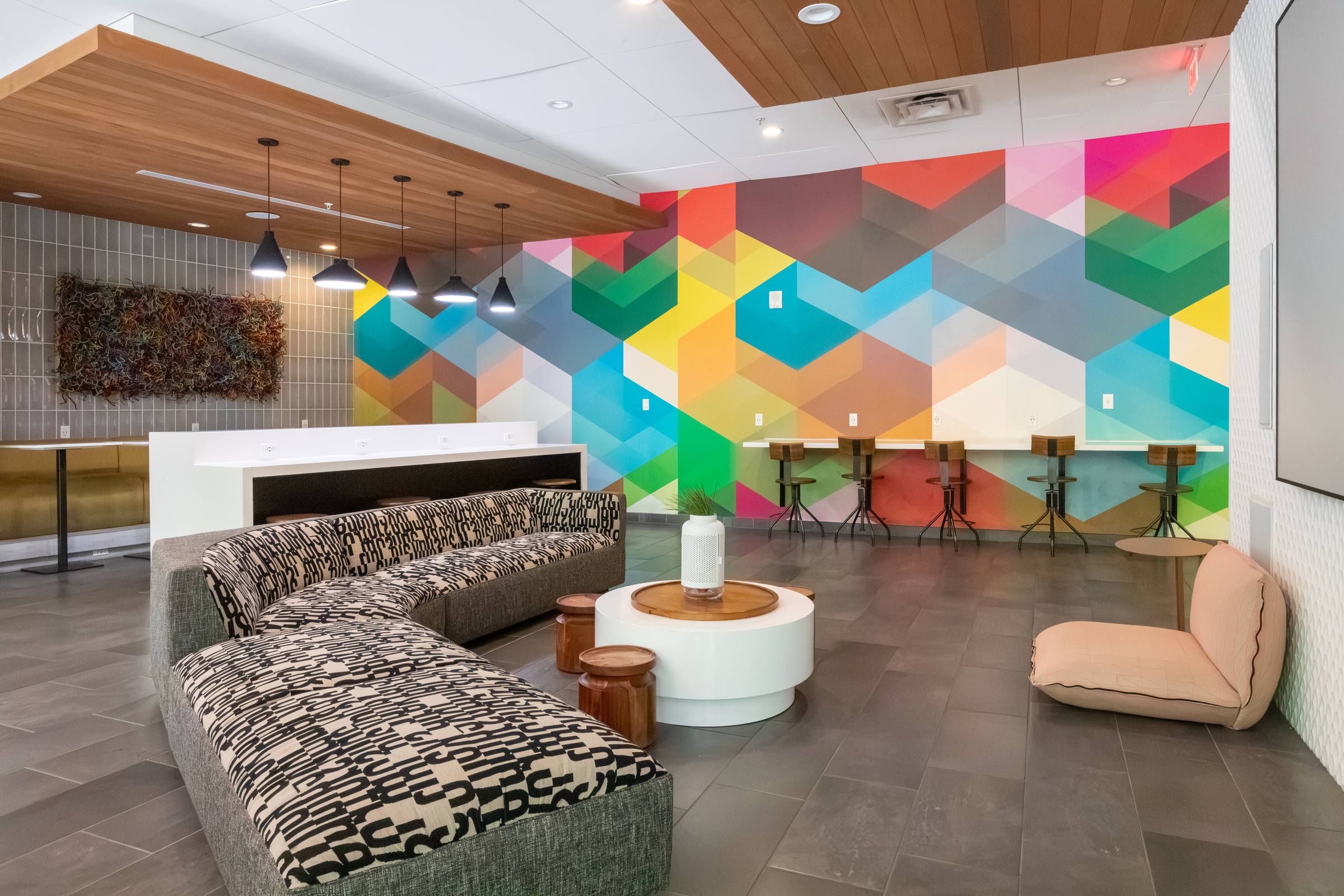 Modern lounge with patterned couches, round tables, bar stools, geometric mural, and hanging lights.